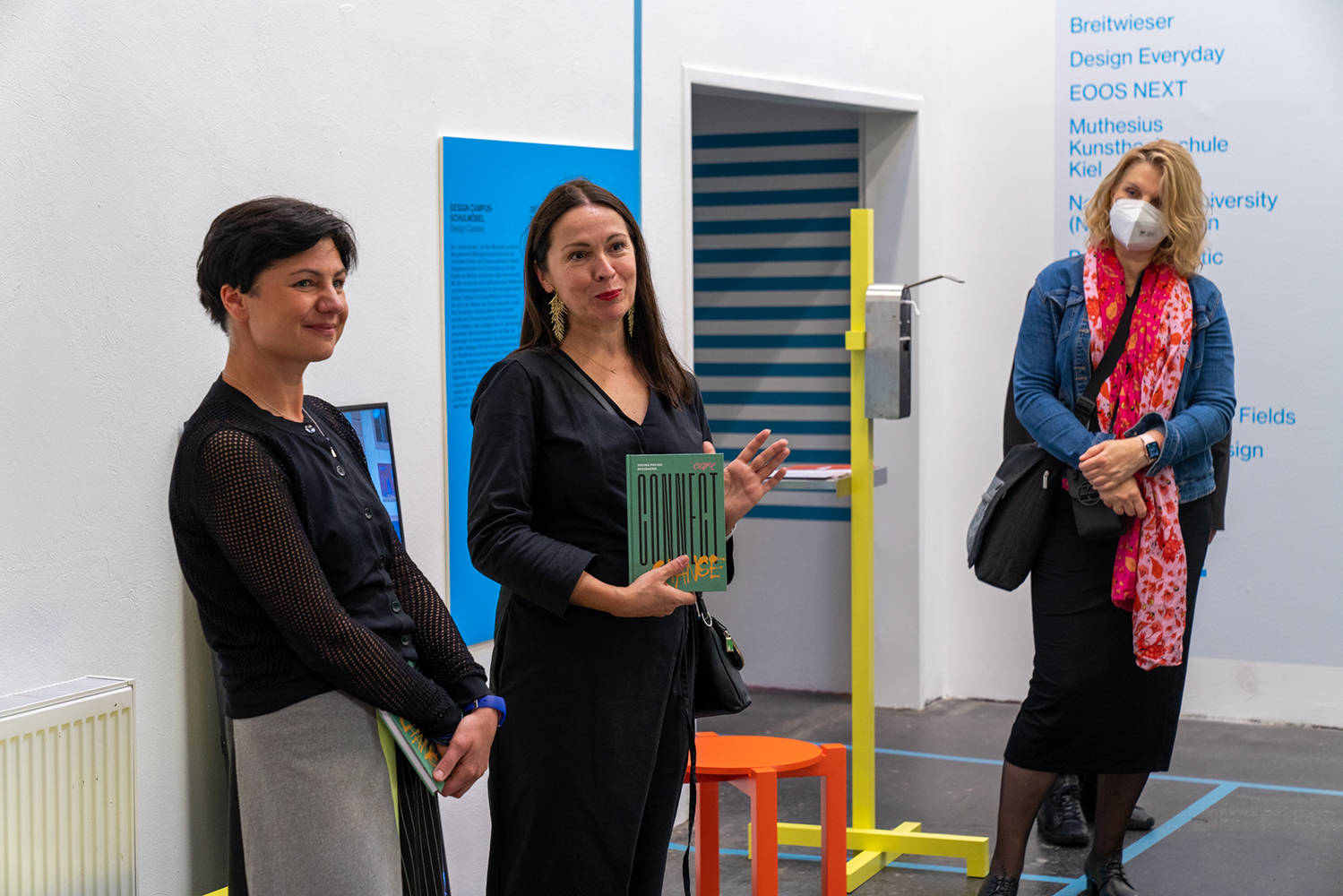 W kadrze trzy kobiety, stojące pośród eksponatów, plansz informacyjnych i napisów umieszczonych błękitnym drukiem bezpośrednio na białej ścianie. Środkową pozycję zajmuje kobieta w czarnej sukience, trzymająca przed sobą zielony katalog z napisem CONNECT i przemawiająca do zebranych
